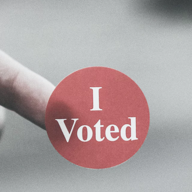 A hand holds a red sticker that reads "I Voted"