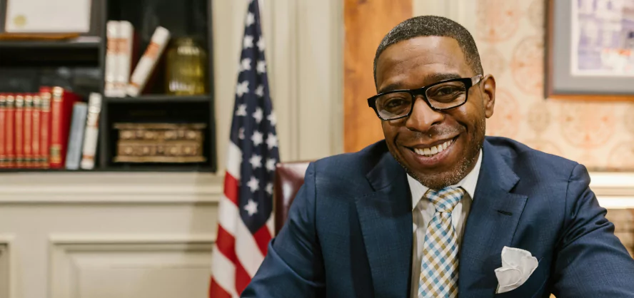 A smiling black man in a suit and tie seated at a desk